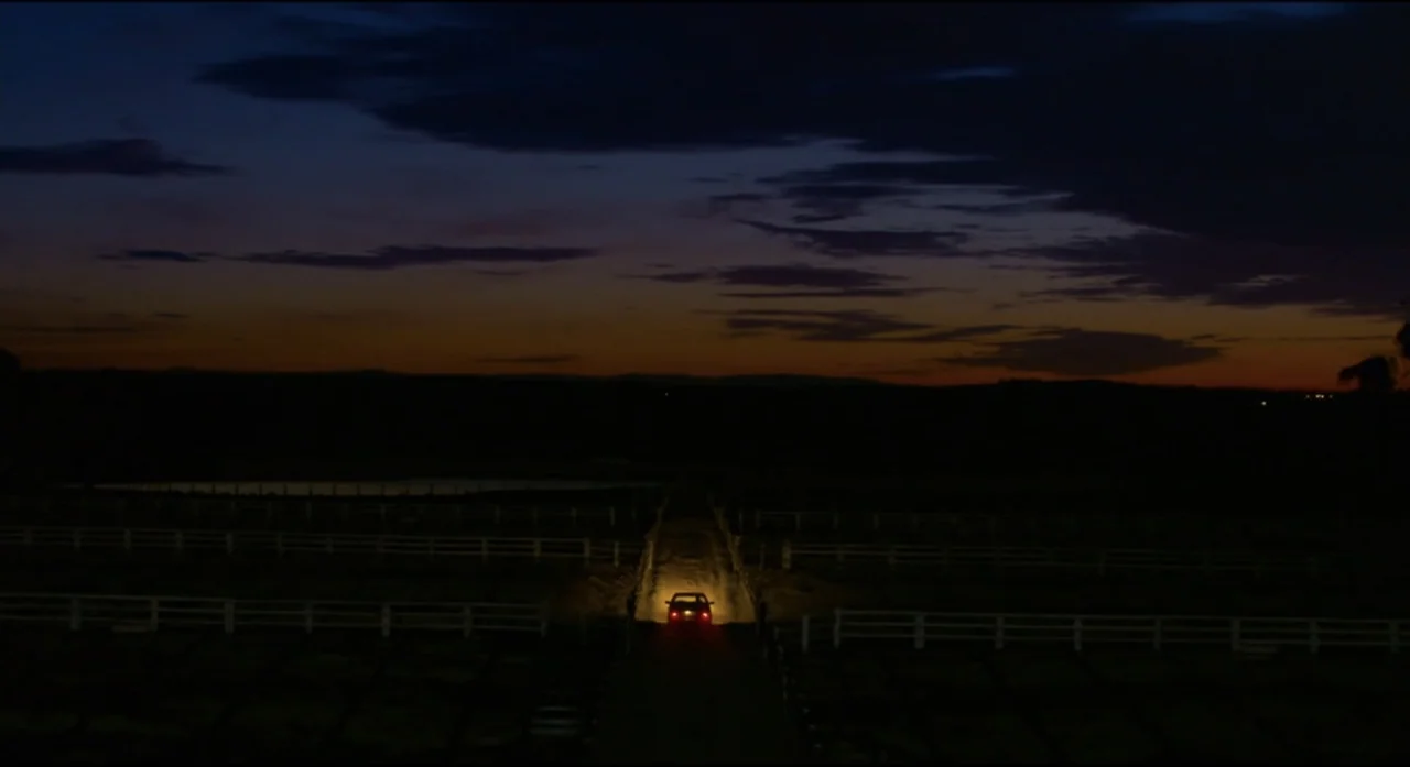 Ride Like a Girl (2019) movie still: wide — A rural landscape at dusk features a long driveway flanked by white wooden fences. A lone car moves…