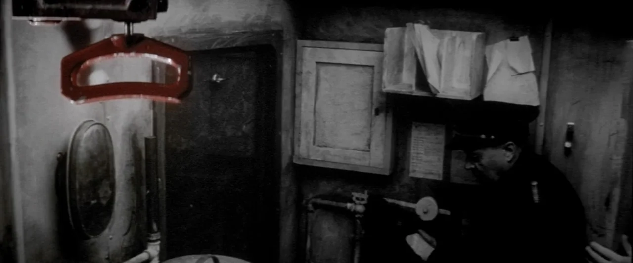Europa (1991) movie still: medium — A dark and confined train cabin features metallic walls and a small storage cabinet. A man in a dark uniform…