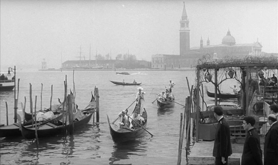 Eva (1962) movie still: wide — A canal view in Venice features a large church dome and bell tower backdrop. Several wooden gondolas carrying…