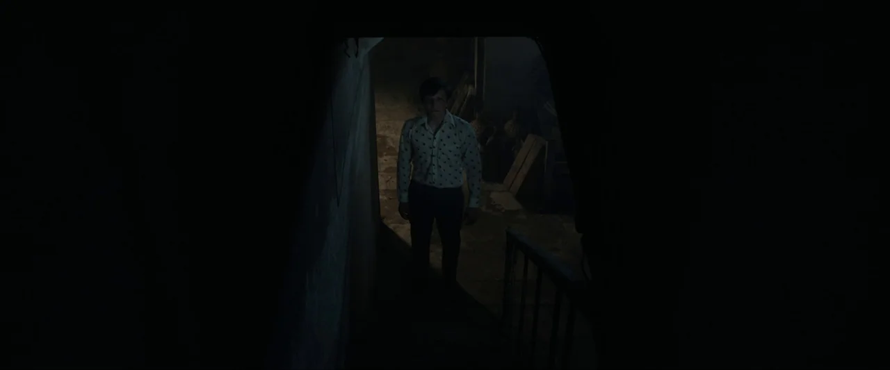 Fréwaka (2024) movie still: fullbody — A dark, cramped interior stairwell is illuminated by a single dim light source. A man in a light patterned…