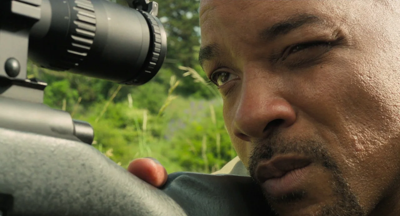 Gemini Man (2019) movie still: closeup — A grassy field with blurred green vegetation serves as the backdrop. A man aims a rifle with a scope…
