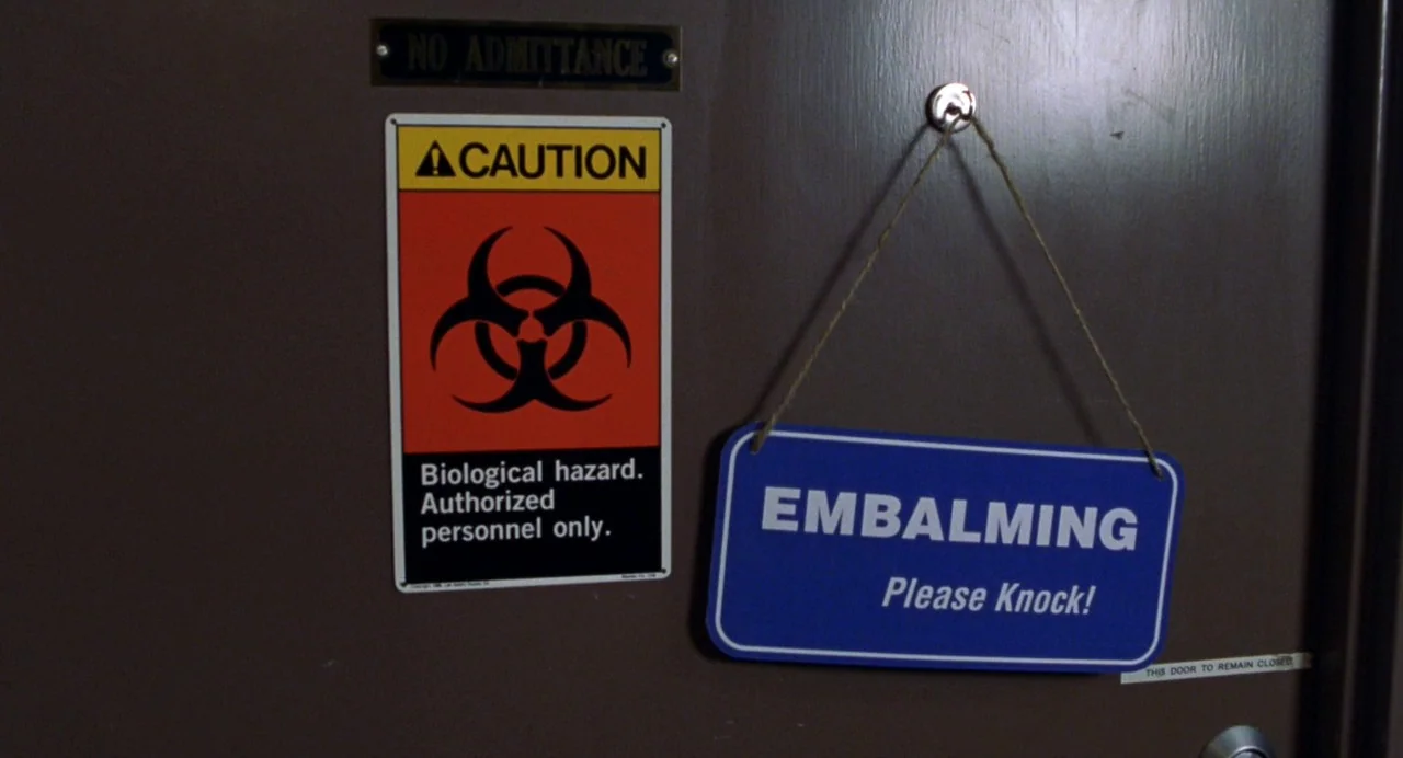 Drop Dead Gorgeous (1999) movie still: closeup — A dark brown wooden door features a biohazard warning sign and an embalming notice. Both signs are…