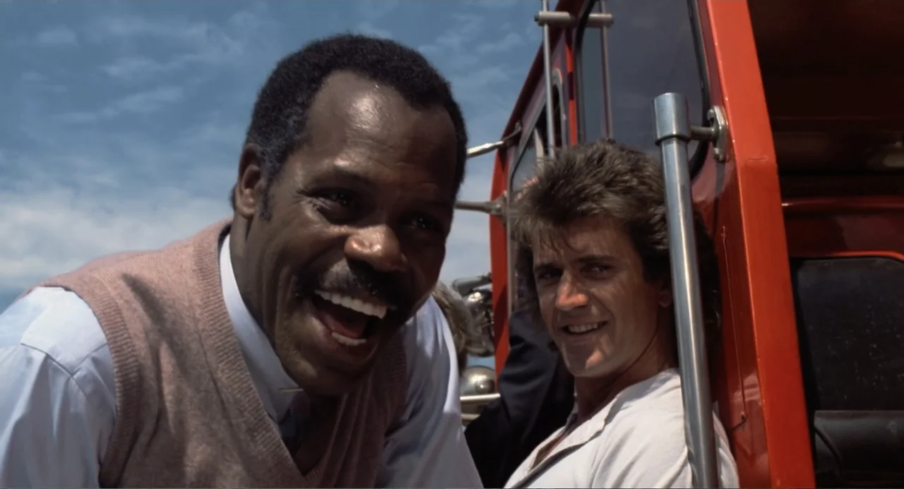 Lethal Weapon (1987) movie still: medium — Two men sit together on the exterior of a bright red fire rescue vehicle. The background shows a clear…
