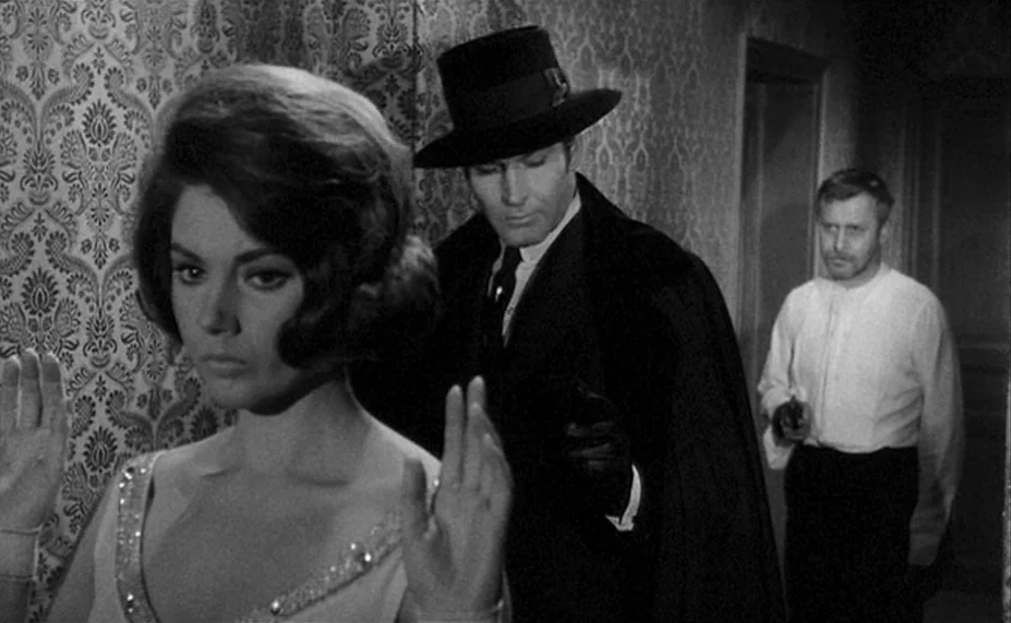 Judex (1966) movie still: medium — A narrow corridor features damask-patterned wallpaper and plain door frames. A man in a hat holds a handgun while…