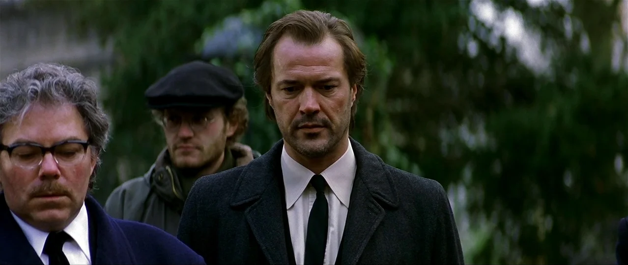 The Lives of Others (2006) movie still: medium — A group of three men stand in a dense green outdoor area with trees. The men wear formal dark coats…