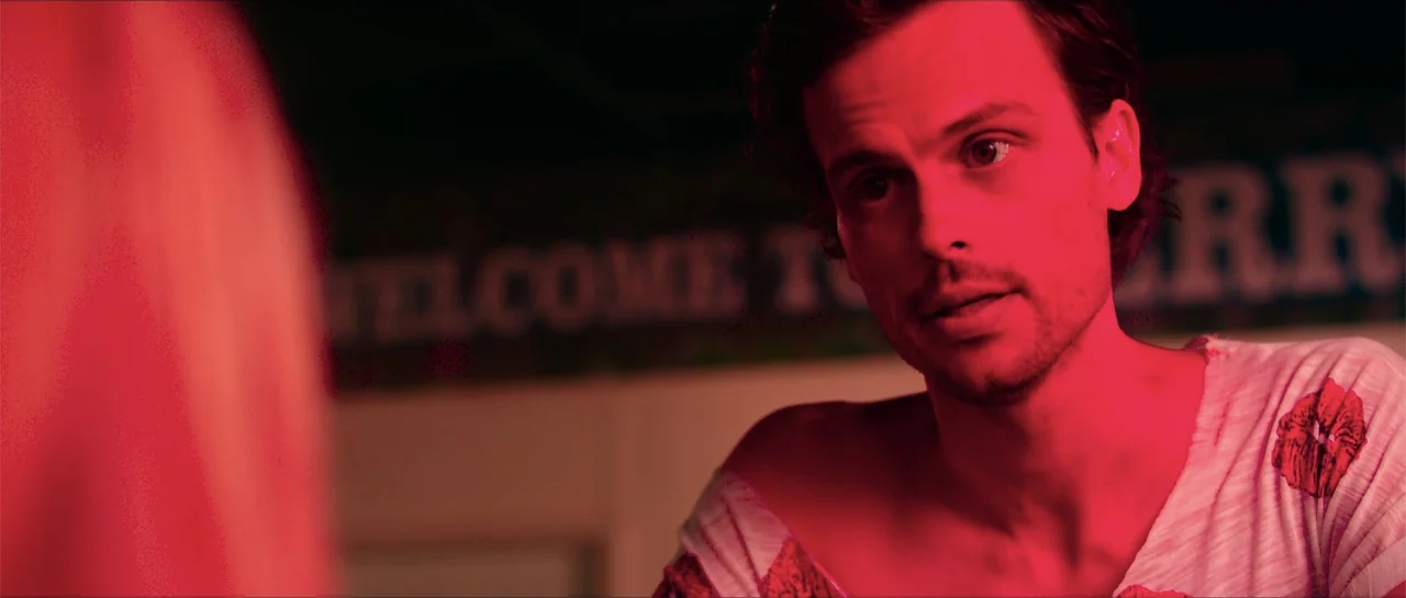 68 Kill (2017) movie still: medium — A dimly lit interior space features a blurred sign in the background. A man sits facing an unseen companion…