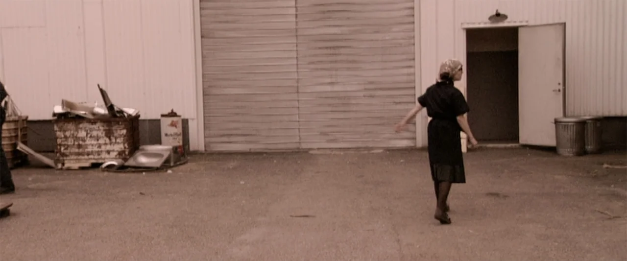 Dancer in the Dark (2000) movie still: fullbody — A paved industrial area features corrugated metal walls and a large, closed sliding door. A woman…