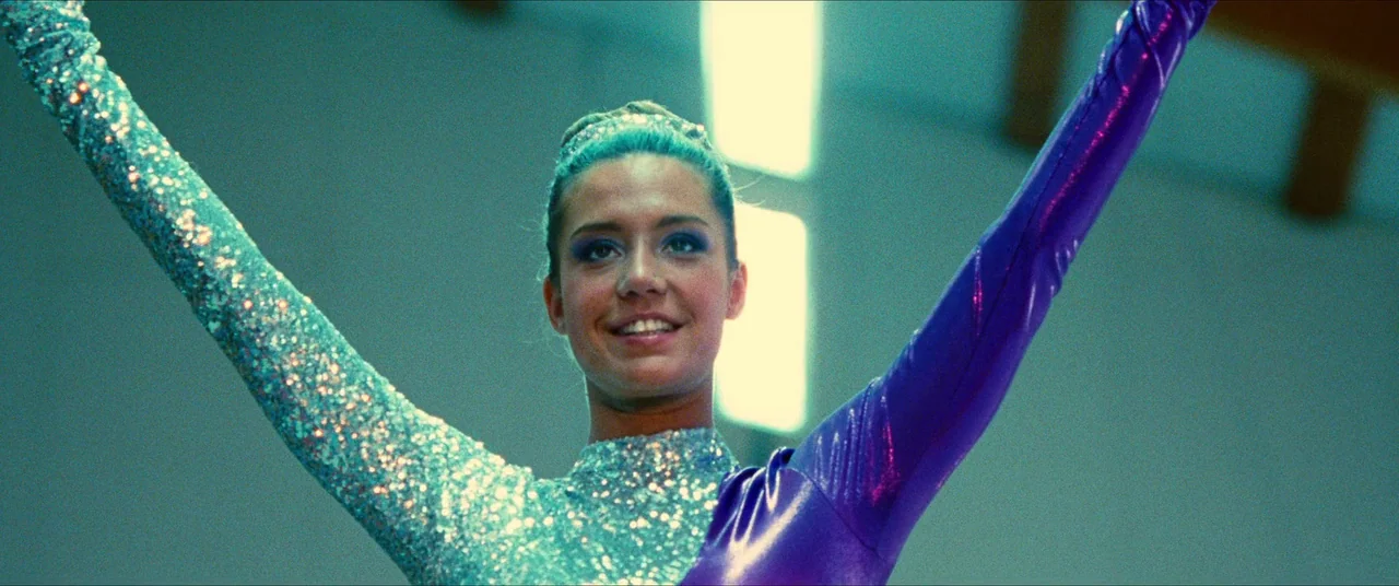 The Five Devils (2022) movie still: medium — A woman in a bright athletic leotard performs in a sterile indoor gymnasium space. She stands with her…