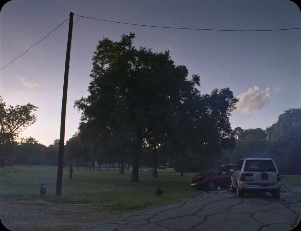 A Ghost Story (2017) movie still: wide — A grassy park area borders a cracked asphalt path at dusk. Two damaged vehicles sit stationary on the…