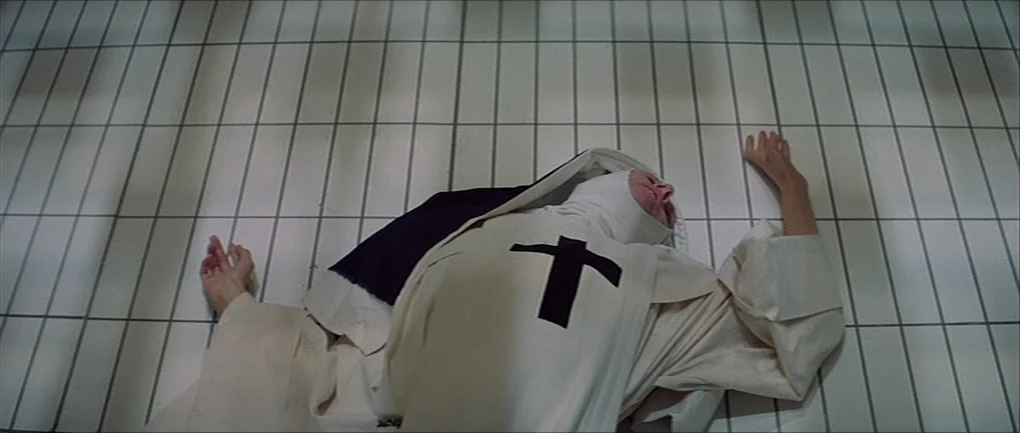 The Devils (1971) movie still: medium — A person wearing a religious habit lies on a stark, white, grid-patterned tile floor. The subject is…