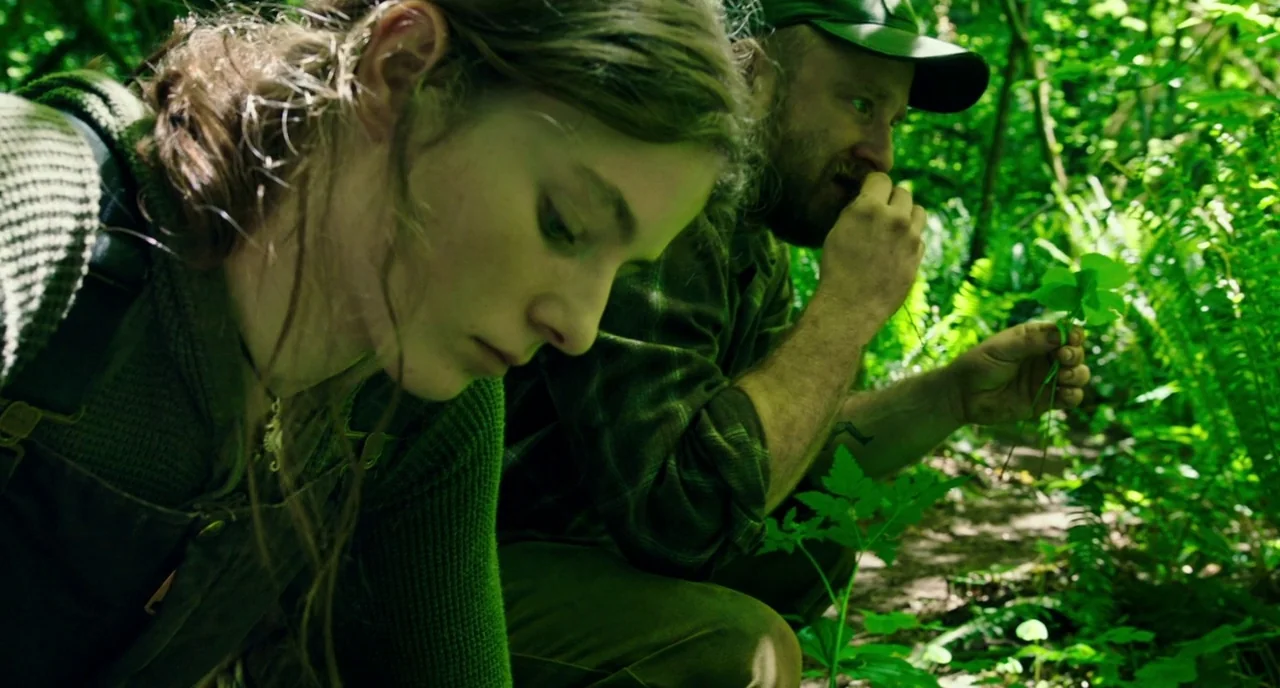 Leave No Trace (2018) movie still: medium — A dense patch of green ferns and wild foliage fills the frame under sunlight. A woman and a bearded man…