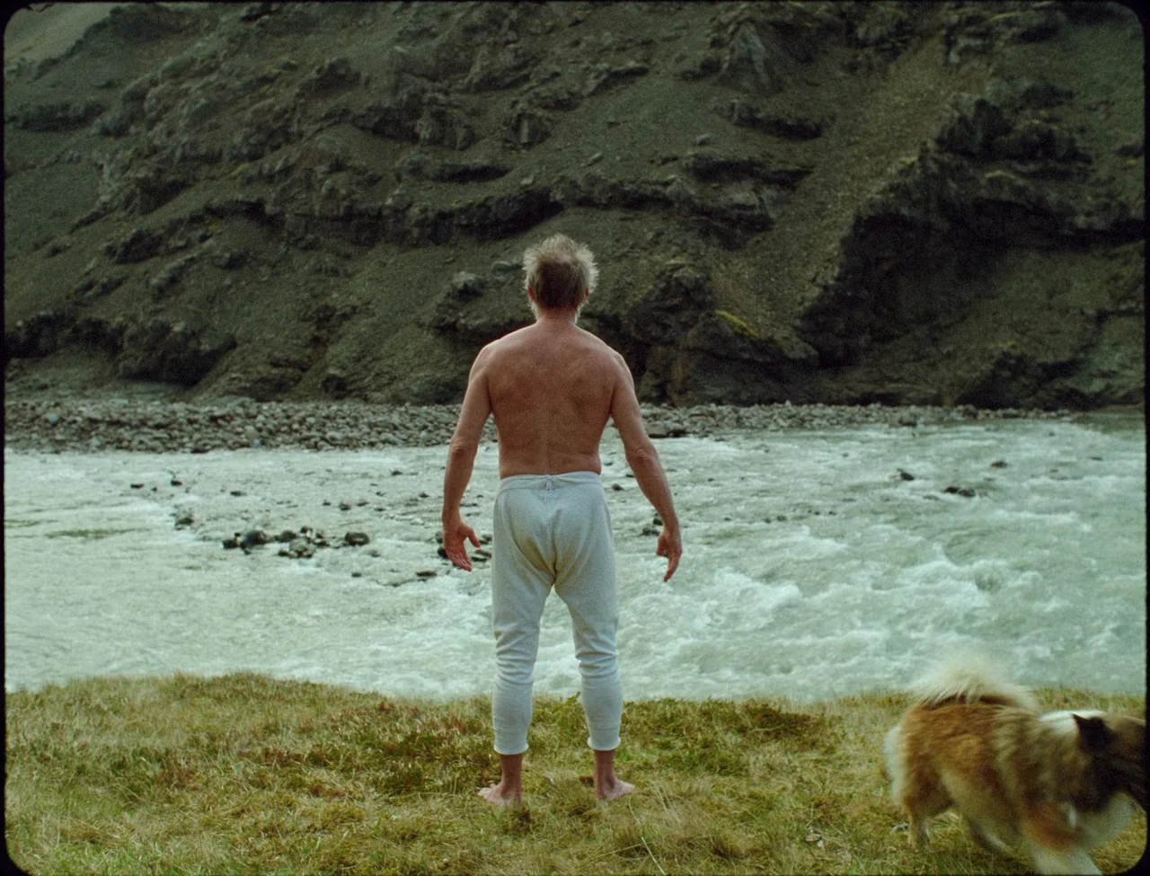 Godland (2022) movie still: fullbody — A grassy bank sits adjacent to a fast-moving, whitewater mountain river with rocky slopes. An elderly man in…