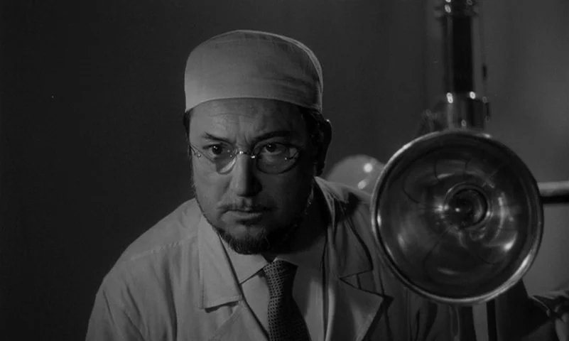 Eyes Without A Face (1960) movie still: closeup — A dimly lit room contains medical equipment including a large lamp. A man in medical attire gazes…