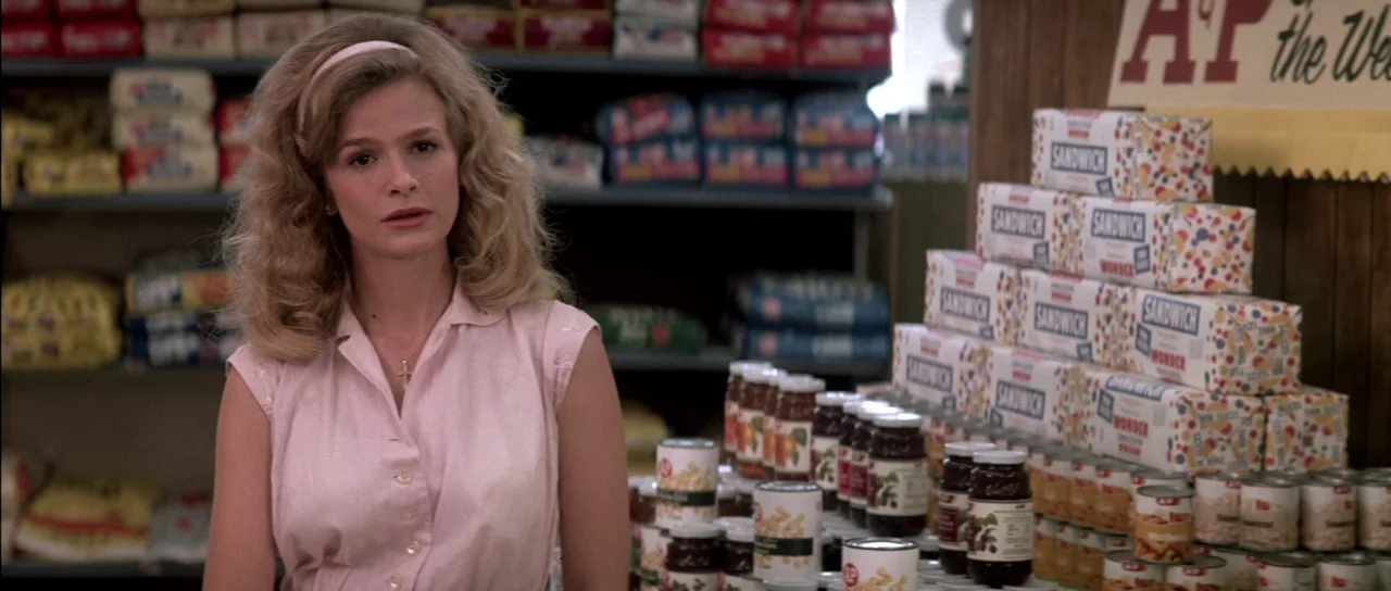 Born On The Fourth of July (1989) movie still: medium — A woman stands in a brightly lit aisle filled with stacked bread and canned goods. She gazes…