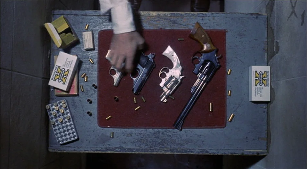 Taxi Driver (1976) movie still: medium — A grey wooden table holds a collection of handguns and boxes of ammunition. A single hand enters the frame…