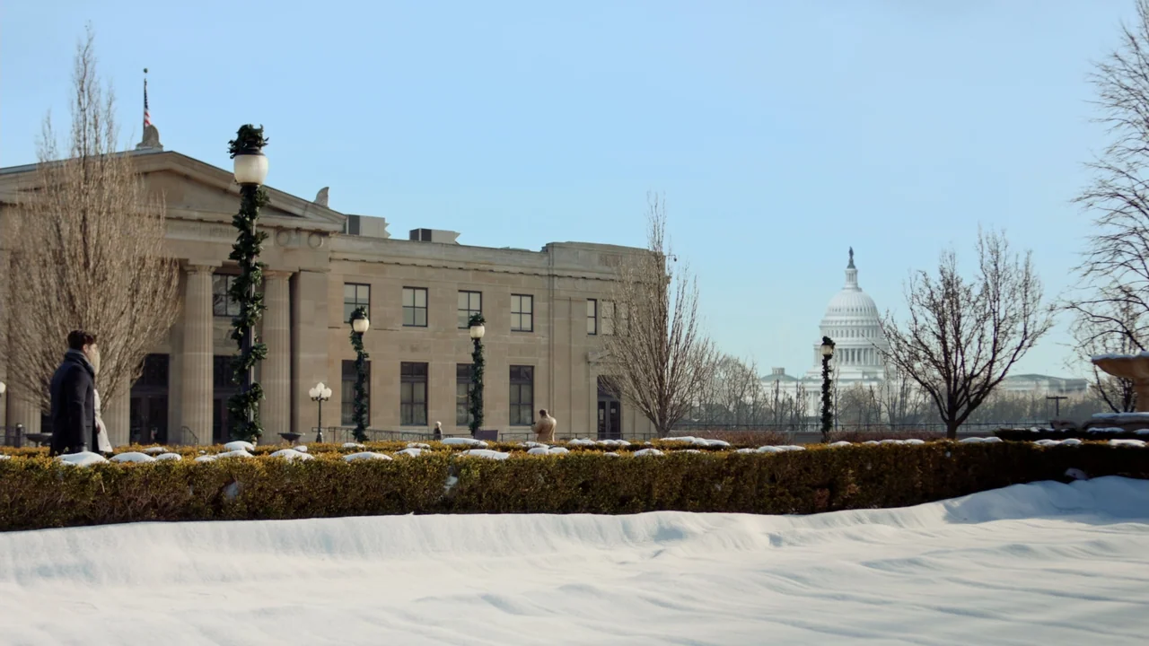 Christmas in Washington (2003) movie still: wide — A snow-covered public park in Washington D.C. features a large stone building facade. A man walks…