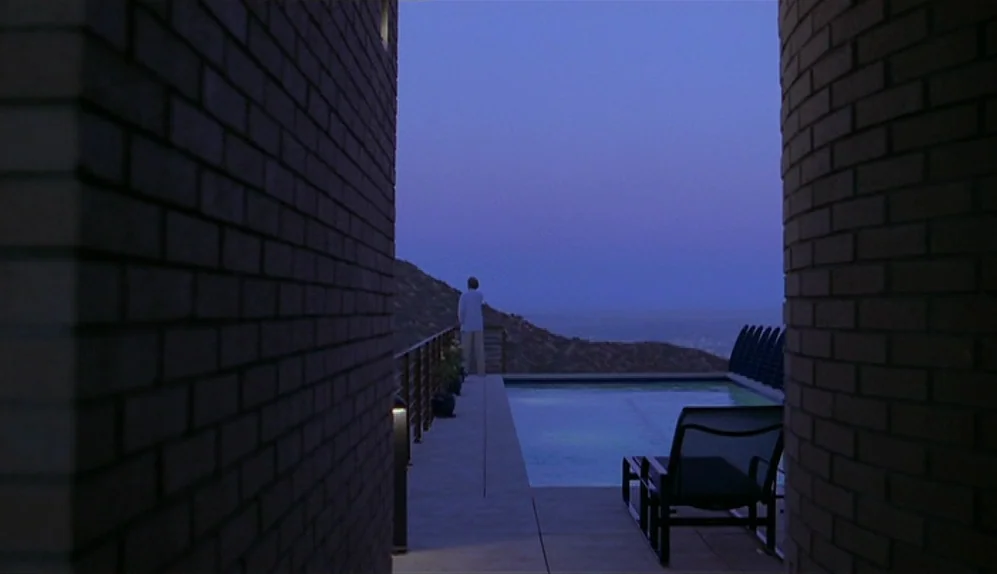 The Limey (1999) movie still: wide — A modern stone terrace overlooks a hillside during the quiet blue hour at twilight. A lone figure stands near…