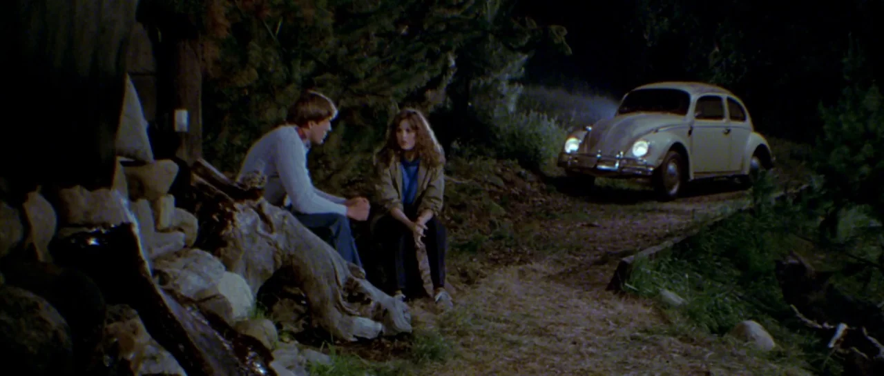 Friday the 13th Part III (1982) movie still: wide — A man and woman sit on a log beside a dirt trail at night. A parked car with illuminated…