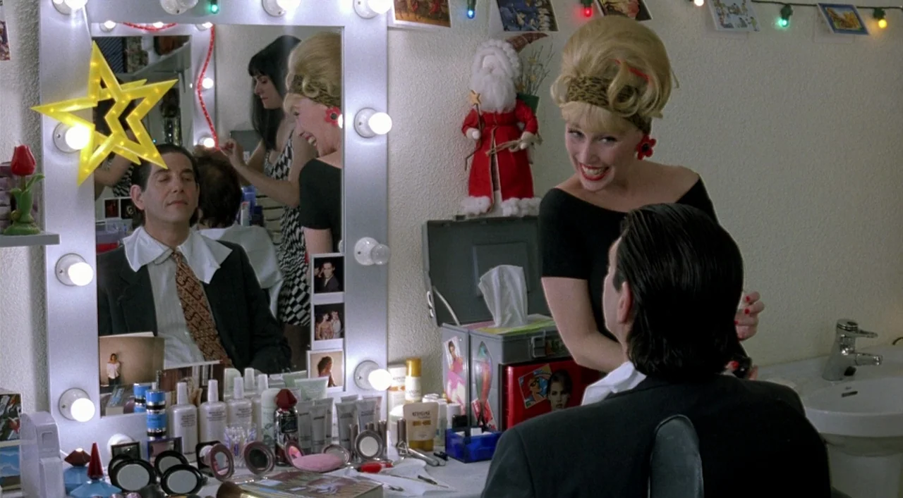 Kika (1993) movie still: medium — A cluttered backstage vanity area is decorated with Christmas lights and a Santa doll. A blonde woman speaks to a…