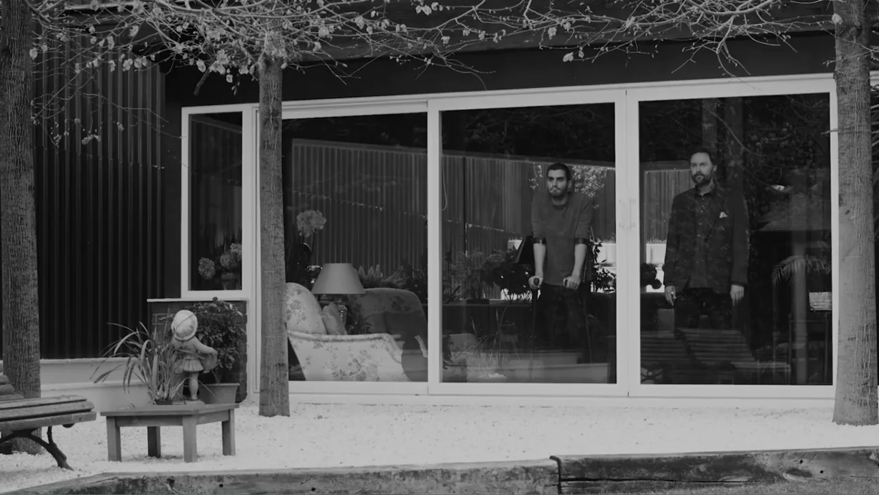 Creditors (2025) movie still: wide — A modern building exterior features large glass windows reflecting the outdoor gravel courtyard. Two men stand…