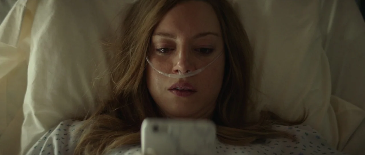 Ingrid Goes West (2017) movie still: closeup — A woman lies in a hospital bed with a nasal cannula. She looks down at a handheld smartphone screen.