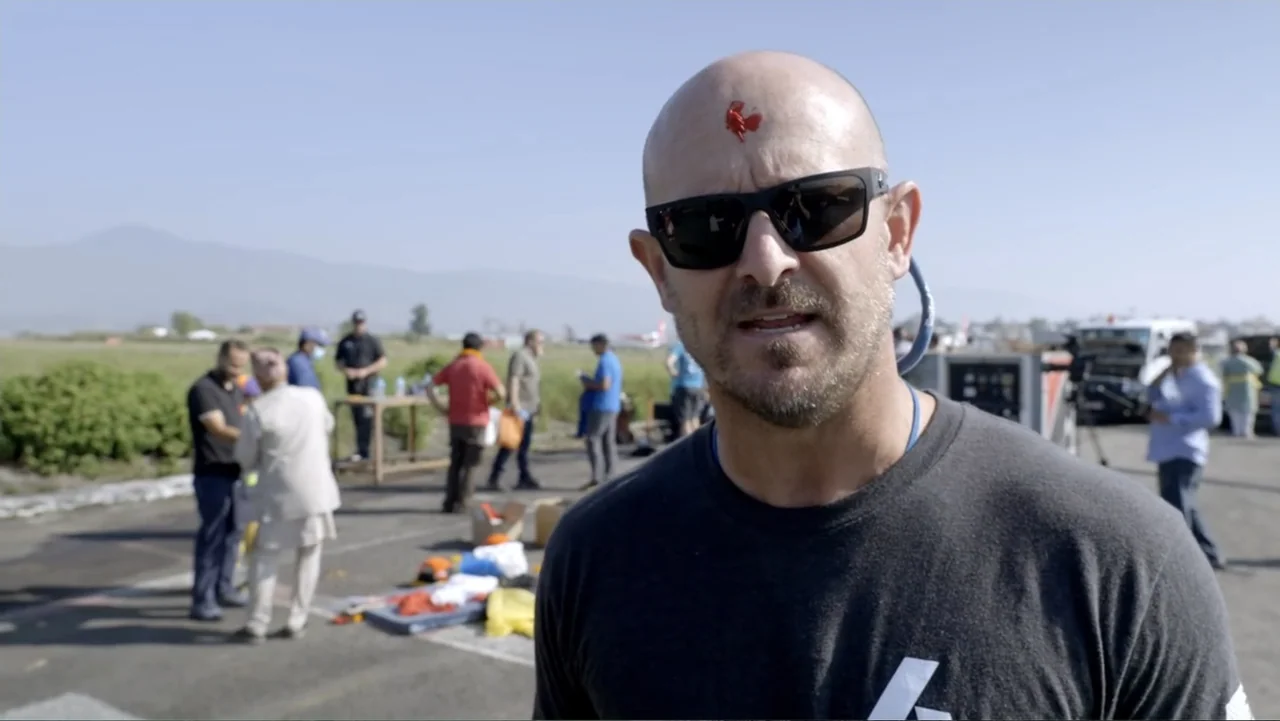 Drop Zone Everest (2023) movie still: medium — An outdoor airport tarmac under a clear sky with several people in the distance. A bald man in a…