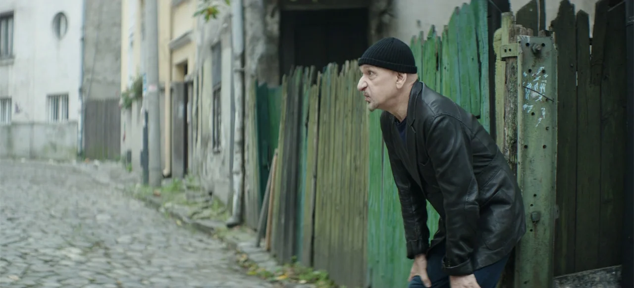 Morir un Poco (1967) movie still: medium — An aged cobblestone alley features a worn, green-painted wooden fence along the right wall. A man leans…