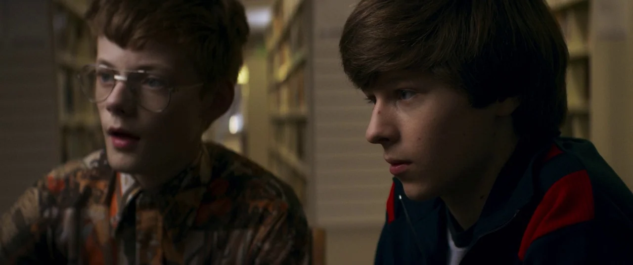Summer of 84 (2018) movie still: medium — Two teenage boys sit together in the aisle of a quiet library with tall shelves. The boys look ahead, one…