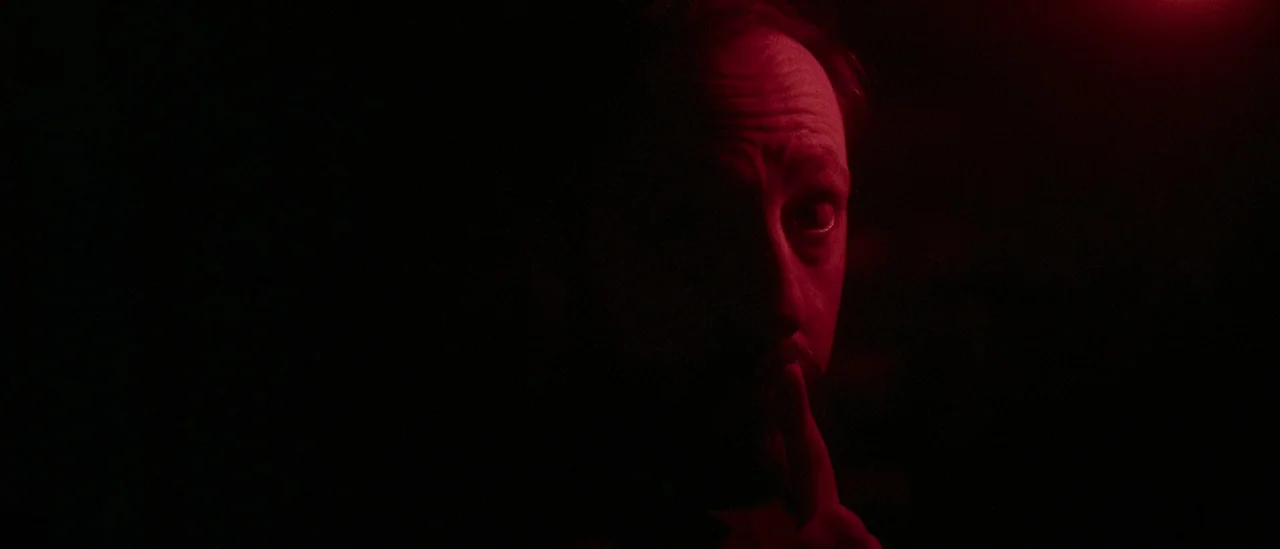 I Trapped The Devil (2019) movie still: closeup — A dimly lit, near-black environment is pierced by intense crimson light. A man with visible…