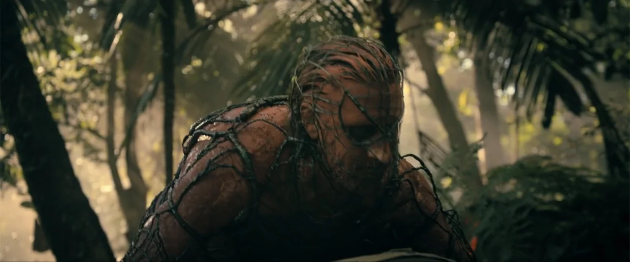Madame Web (2024) movie still: medium — A dense tropical thicket features tall palm trees and thick undergrowth in soft light. A mud-covered man…