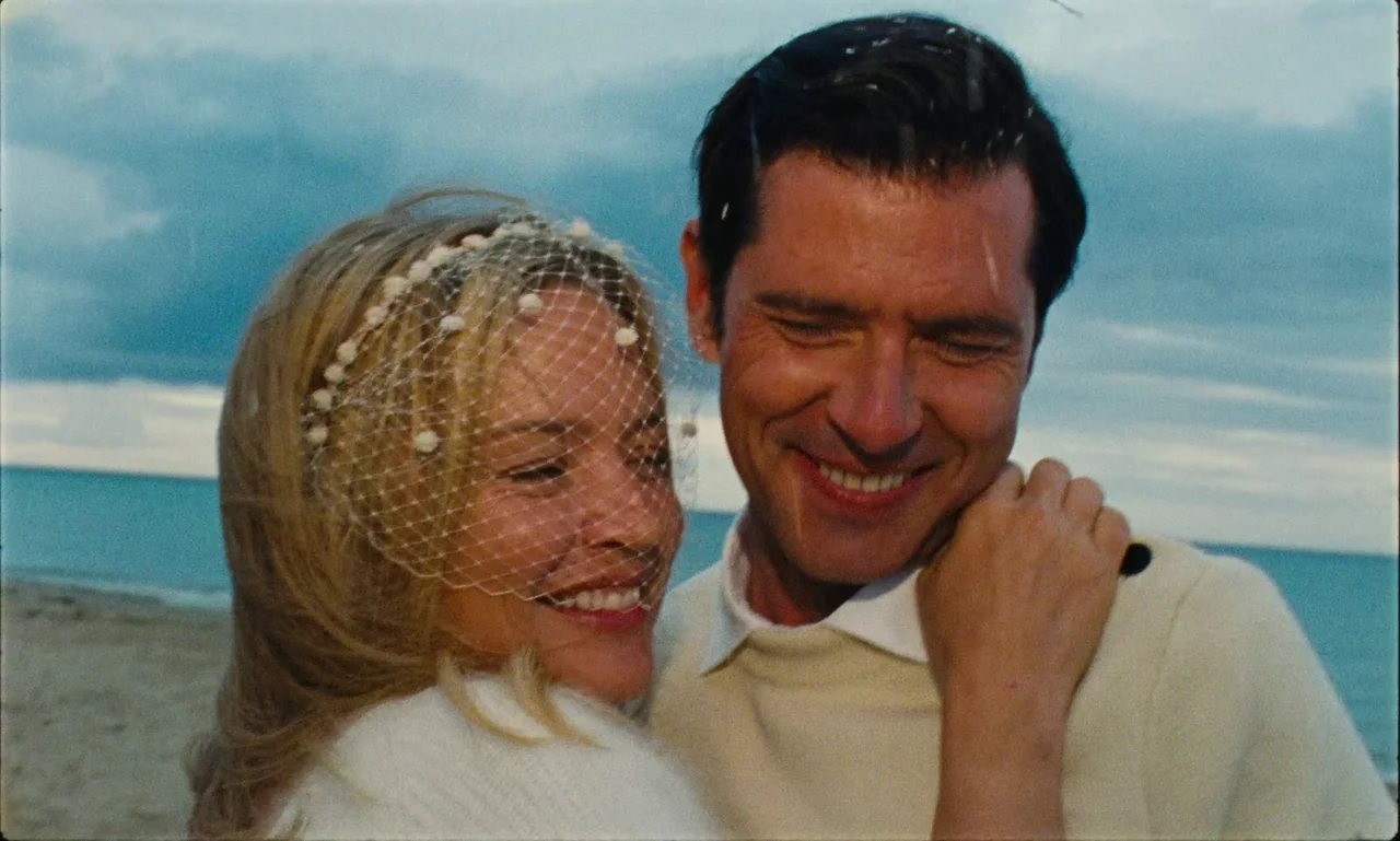 Just The Two Of Us (2023) movie still: medium — A man and woman embrace on a sandy shore under a cloudy sky. The ocean is visible in the background…