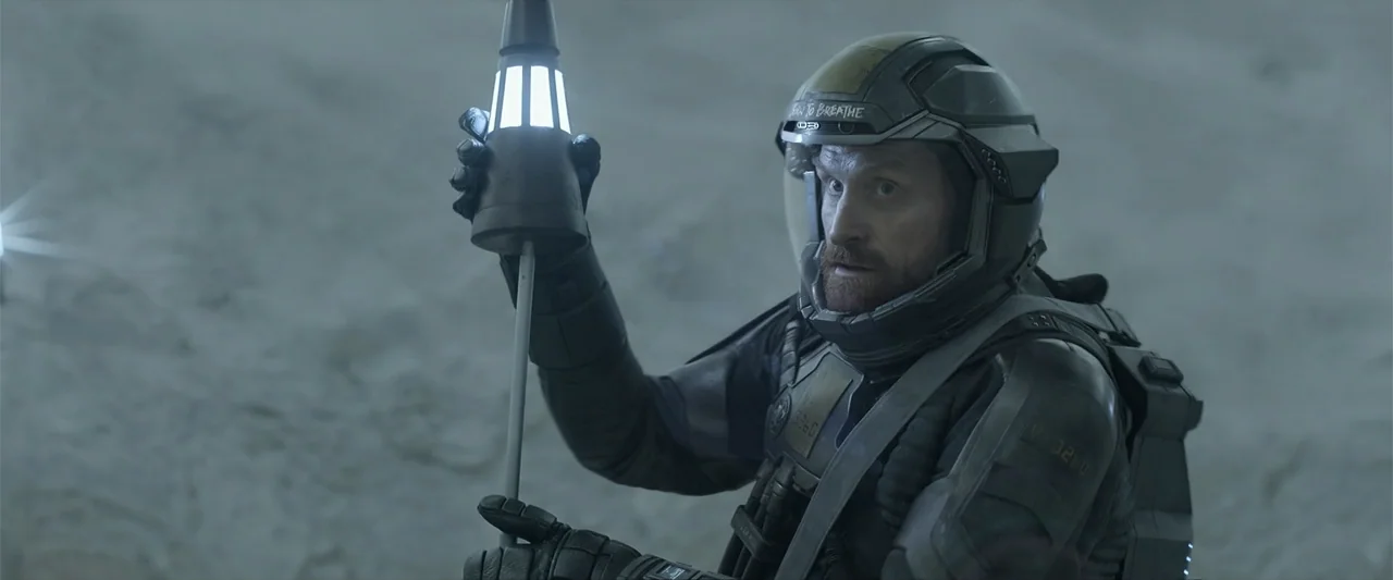DISTANT (2026) movie still: medium — A desolate and rocky landscape at night under dim atmospheric light. A space-suited man holds a glowing rod…