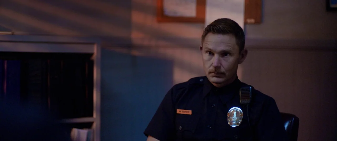 Blindfire (2020) movie still: medium — A dimly lit room features a bulletin board on the back wall and shadows. A police officer in uniform sits in…