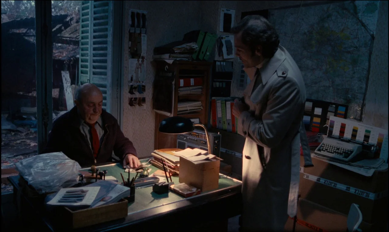 Serie Noire (1979) movie still: medium — A cramped, dimly lit office contains stacks of file folders and a vintage typewriter. An older man sits at…