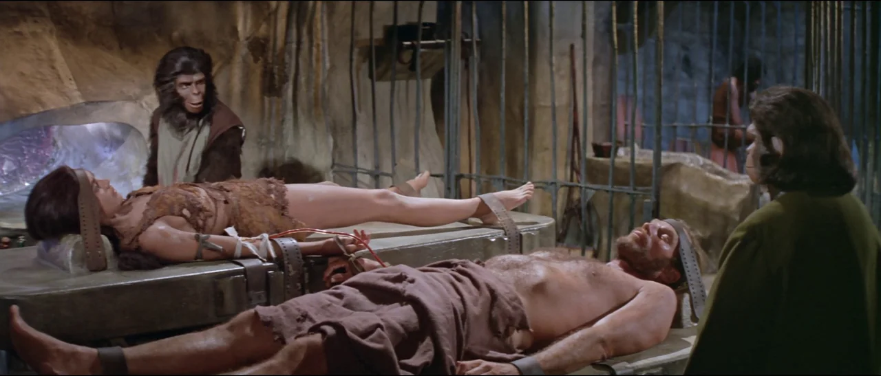 Planet of the Apes (1968) (1968) movie still: medium — A stone-walled underground room with metal cages and surgical tables. Two humans lie…