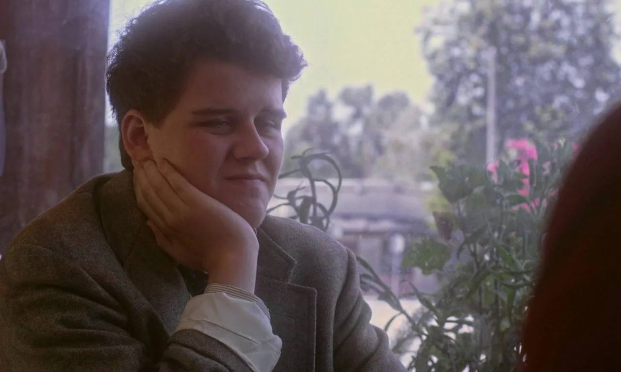 Ham on Rye (2019) movie still: medium — A man sits indoors near a window with blurred foliage visible in the background. He rests his face on his…