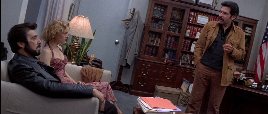 Carlito’s Way (1993) movie still: medium — A well-lit room contains a mahogany bookshelf, a desk, and a seating area. A man and woman sit on a sofa…