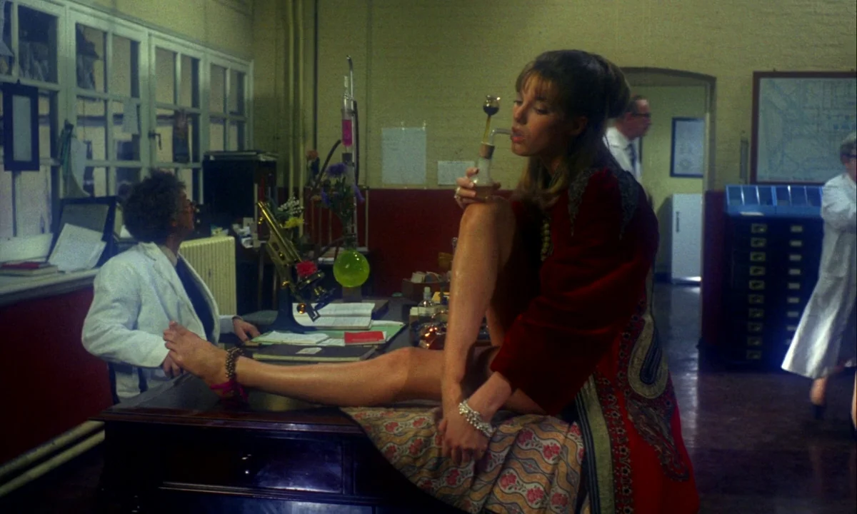 Wonderwall (1968) movie still: medium — A cluttered laboratory features large multi-paned windows and a dark wood desk. A woman sits on the desk…
