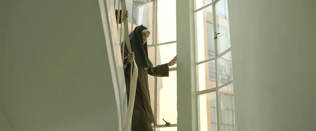 Divine Intervention (2026) movie still: medium — A white-walled interior stairwell features a large window frame and a metal railing. A woman…