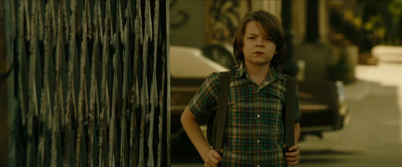 Wonderstruck (2017) movie still: medium — A suburban sidewalk is visible with a metal gate partially obscuring the left frame. A young boy wearing a…