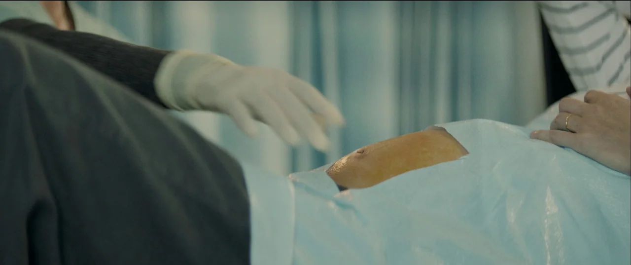 Birth/Rebirth (2023) movie still: closeup — A medical examination room features light blue paper drapes covering a patient's abdomen. A person's…