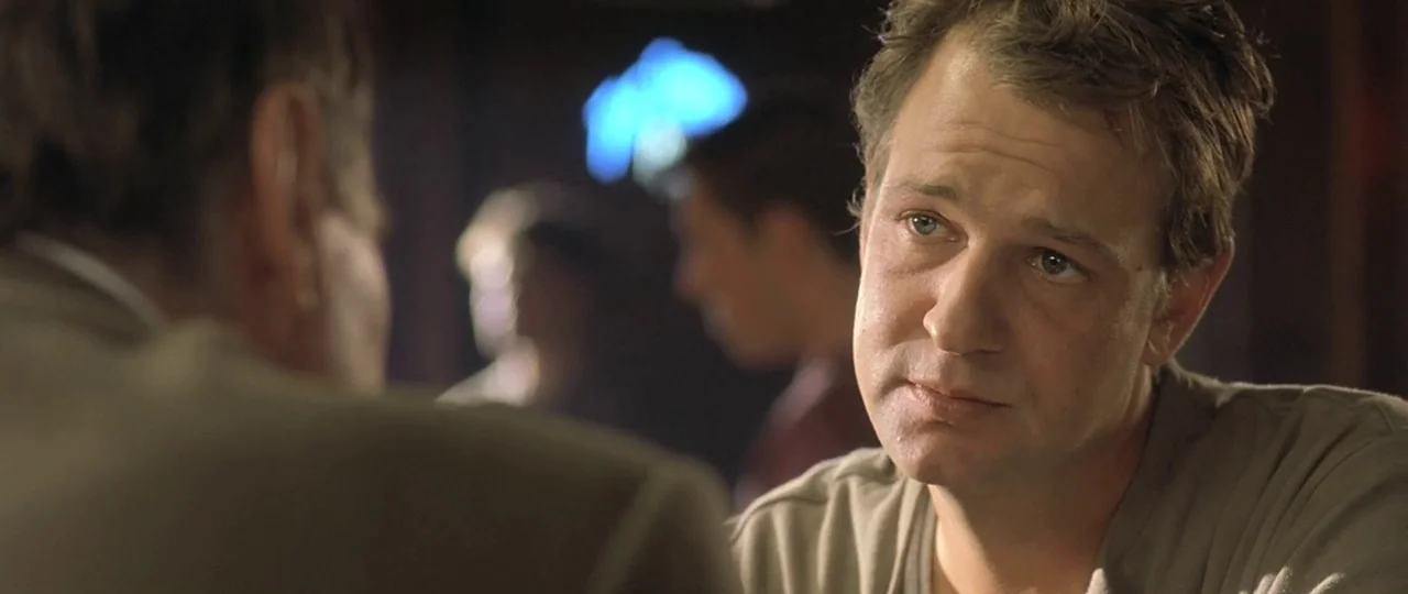 In the Bedroom (2001) movie still: medium — A dimly lit drinking establishment features patrons blurred in the dark background. A man sits at a…