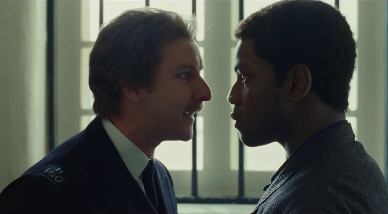 Red, White & Blue (2020) movie still: medium — An interior space with barred windows featuring a man in a police uniform. He shouts inches from the…