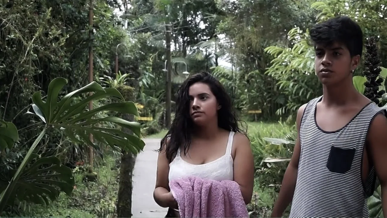 El Duende Sátiro (2016) movie still: medium — A paved walkway winds through a dense tropical garden with large monstera leaves. A woman and a young…