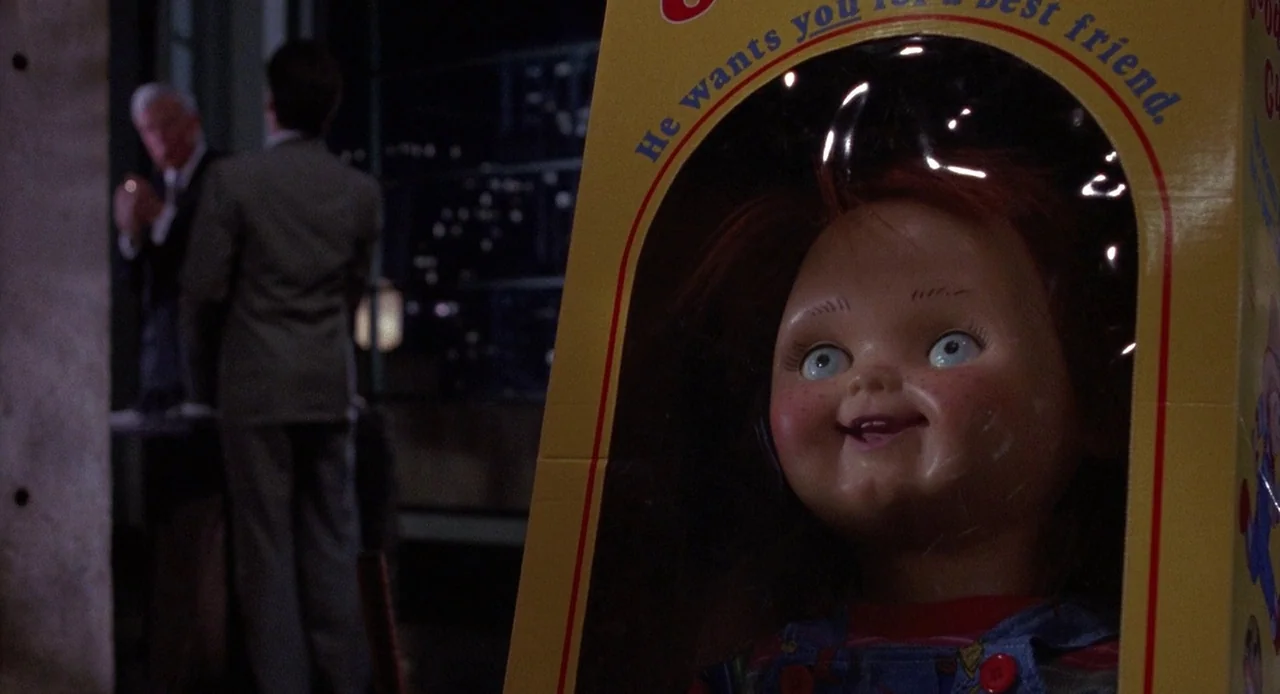 Child’s Play 3 (1991) movie still: closeup — A close-up of a doll inside a yellow cardboard retail box with plastic shielding. In the blurred…