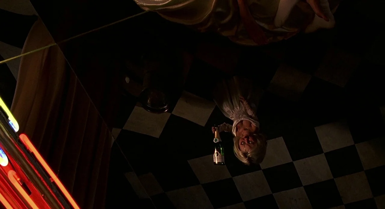 Bride of Chucky (1998) movie still: wide — A dimly lit room features a black and white checkered floor and neon lighting. A small doll dressed in a…