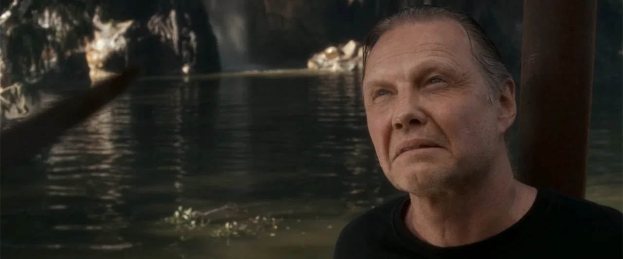 Anaconda (2025) movie still: medium — A dark river flows before a waterfall in a natural outdoor environment. An older man in a black shirt stands…