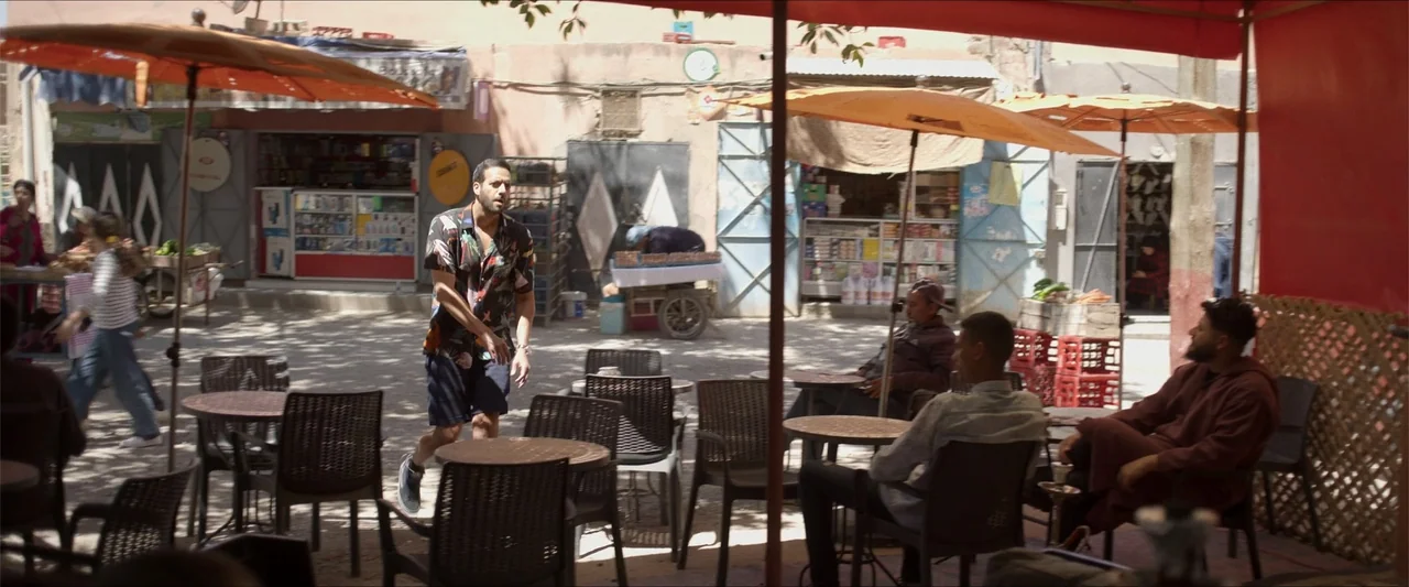 An Endless Wedding (2025) movie still: medium — An outdoor cafe with orange umbrellas and black chairs situated in a town plaza. A man walks through…