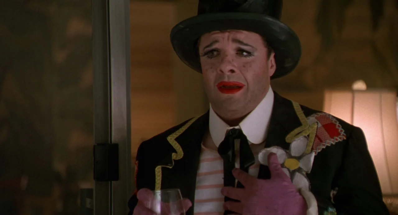 The Birdcage (1996) movie still: closeup — A person wears formal clown attire including a top hat and a floral-embellished jacket. The individual is…