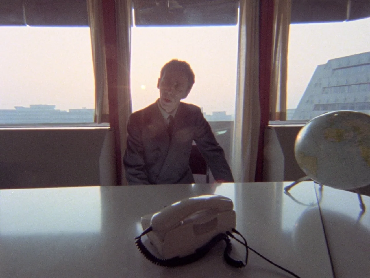 Decoder (1984) movie still: medium — A high-rise office space features a wide window overlooking a sprawling cityscape at dusk. A man sits at a…